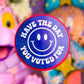 A close-up of a round blue glitter lollipop or badge on a stick, featuring a white smiley face and the text 'HAVE THE DAY YOU VOTED FOR' in white. The background is a colorful, blurred scene with plush toys and decorative items.