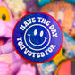 A close-up of a round blue glitter lollipop or sticker with a white smiley face and the text 'HAVE THE DAY YOU VOTED FOR' in white, held in front of a colorful, blurred background of toys and accessories.