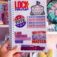 A hand holds a white spiral-bound notebook covered in political stickers with slogans like 'LOCK HIM UP', 'VOTE', and 'I AM WOMAN WATCH ME VOTE', against a pink decorated room background.