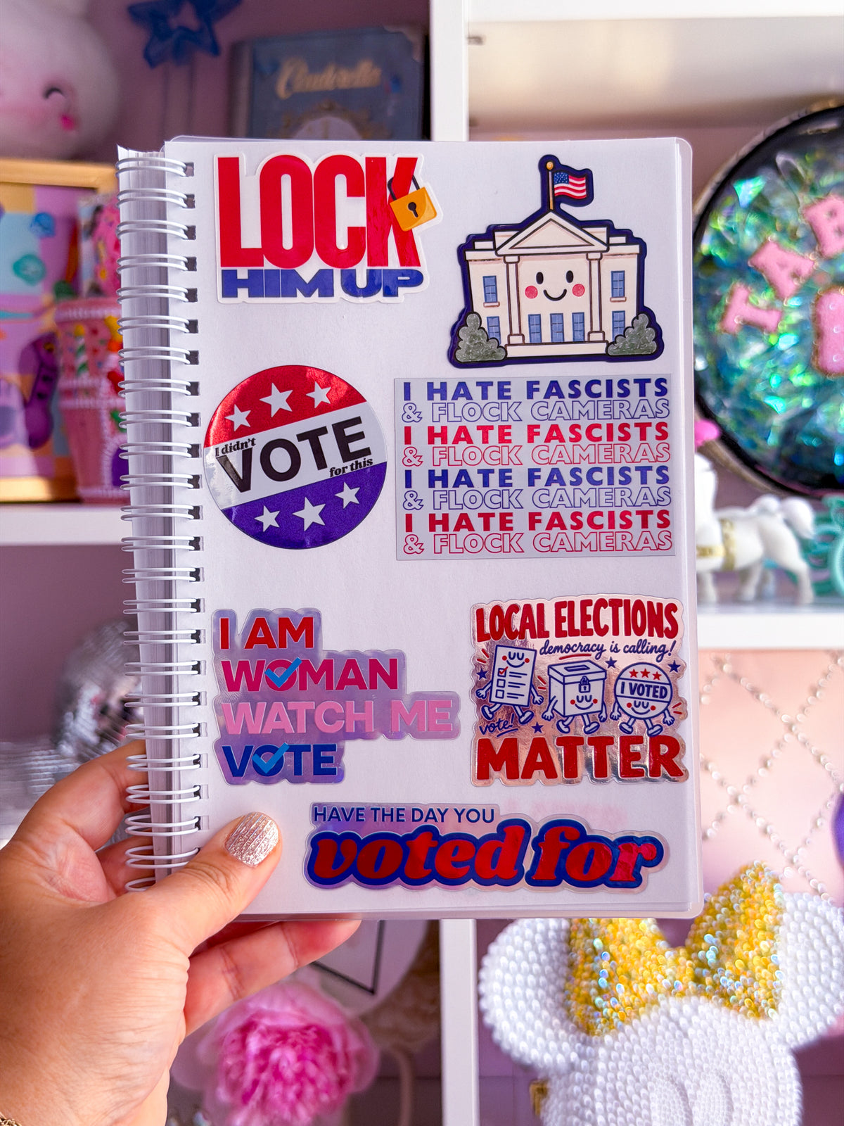 A hand holds a white spiral-bound notebook covered in political stickers with slogans like 'LOCK HIM UP', 'VOTE', and 'I AM WOMAN WATCH ME VOTE', against a pink decorated room background.