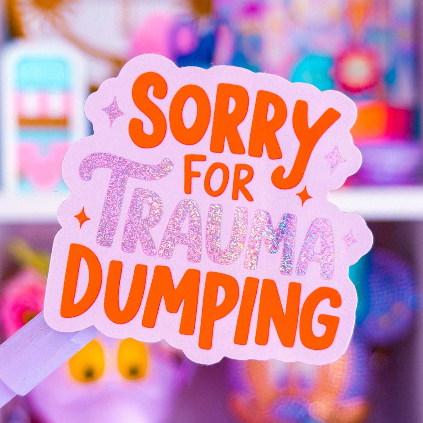 A close-up of a die-cut sticker held in front of a blurred shelf background. The sticker has a light pink border and reads 'SORRY FOR TRAUMA DUMPING' in orange and glittery pink lettering.