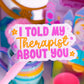A close-up of a pink sign on a stick with the text 'I TOLD MY THERAPIST ABOUT YOU' in pink and orange lettering, surrounded by colorful toy-like objects in a blurred background.