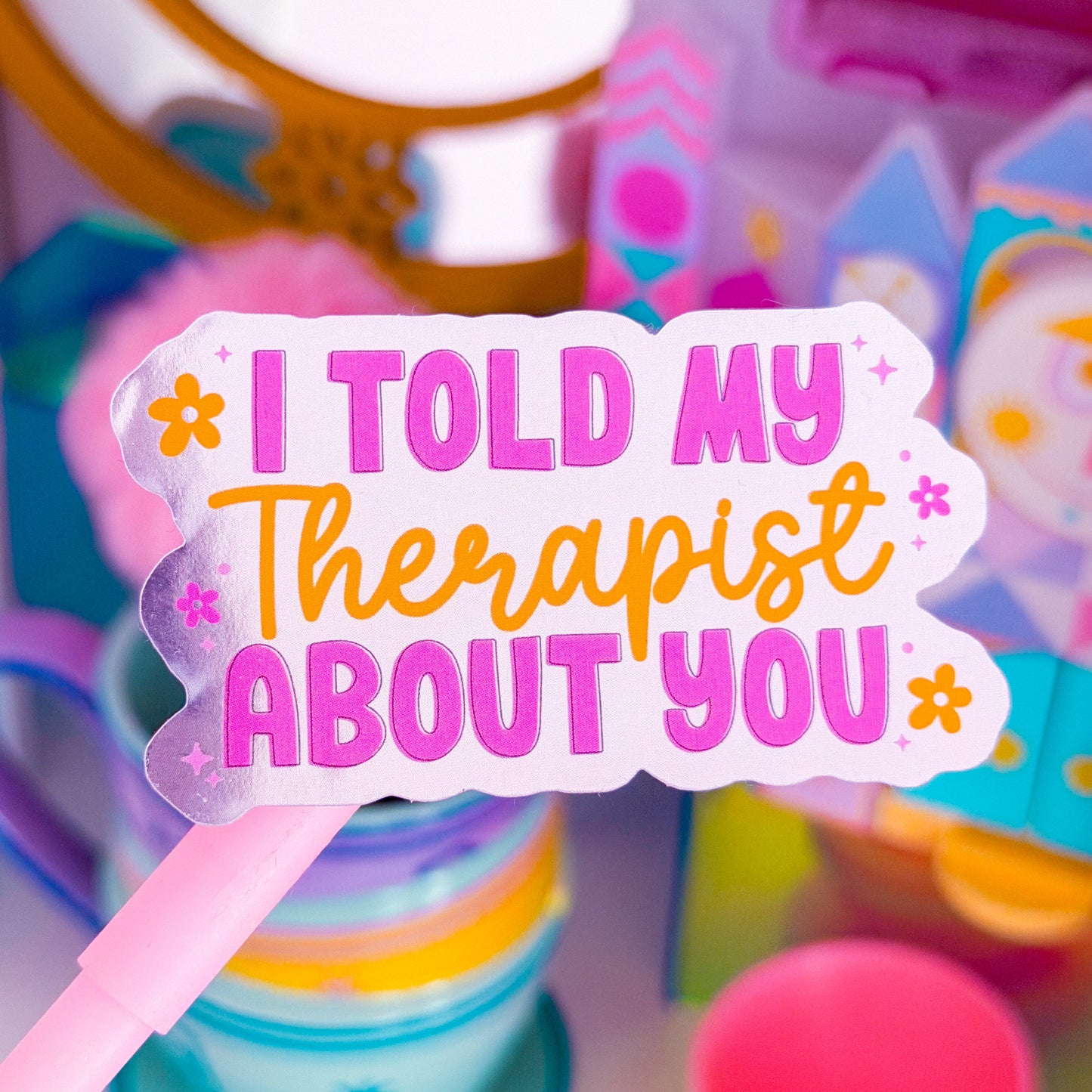 A close-up of a pink sign on a stick with the text 'I TOLD MY THERAPIST ABOUT YOU' in pink and orange lettering, surrounded by colorful toy-like objects in a blurred background.
