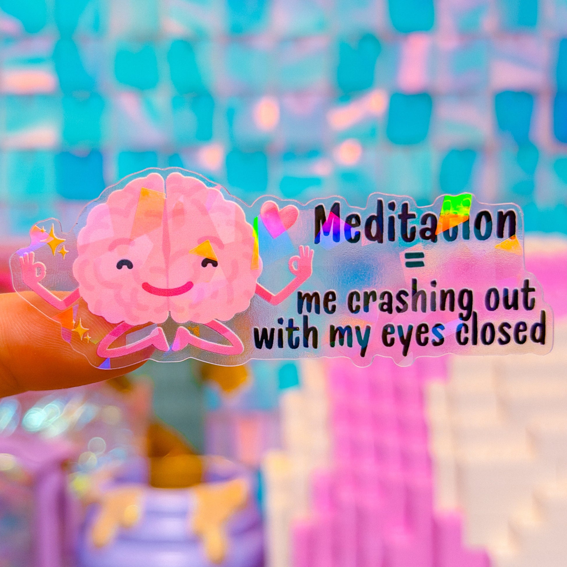 Pink cartoon brain sticker with text 'Meditation = me crashing out with my eyes closed' on a colorful blurred background.